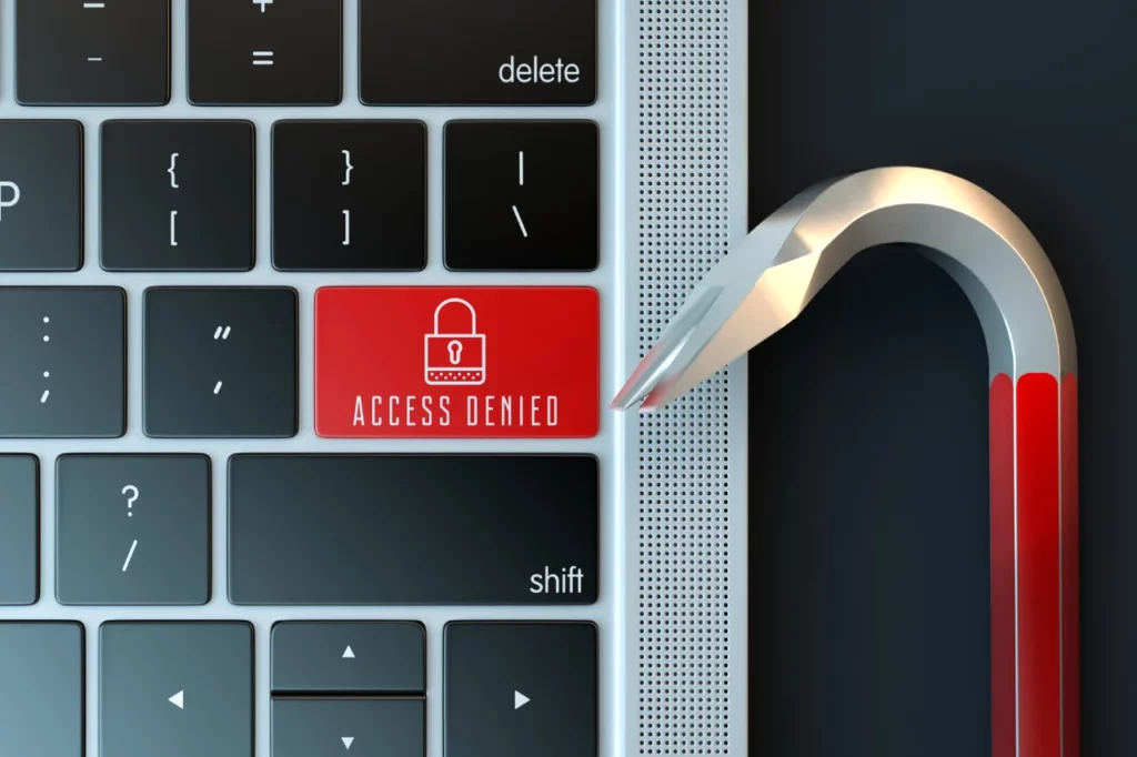 Et tastatur med en stor rød tast, hvor der står "ACCESS DENIED" sammen med et ikon af en lås. Ved siden af tastaturet er der en brækstang, der symboliserer forsøg på at bryde ind eller hacking. Dette billede illustrerer konceptet af cybersikkerhed og beskyttelse mod uautoriseret adgang.